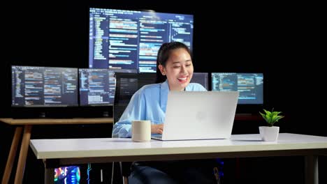 asian female programmer having a meeting by a laptop while writing code on multiple monitors showing database on terminal window desktops in the office