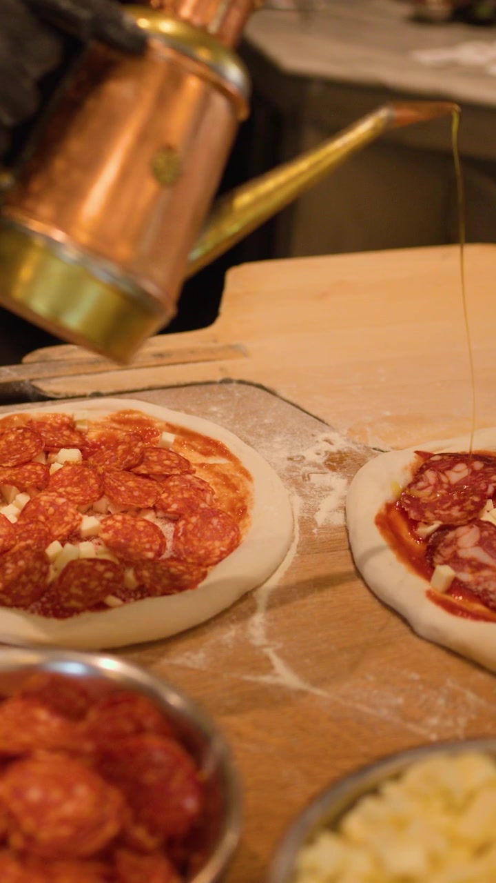 Chef con guanti neri aggiunge pepperoni e olio d'oliva a pizze fatte a mano su un bancone di legno.