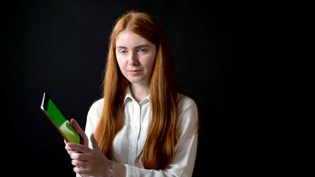 Young ginger woman in white shirt holding tablet with chroma key and showing thumbs up, isolated on black background