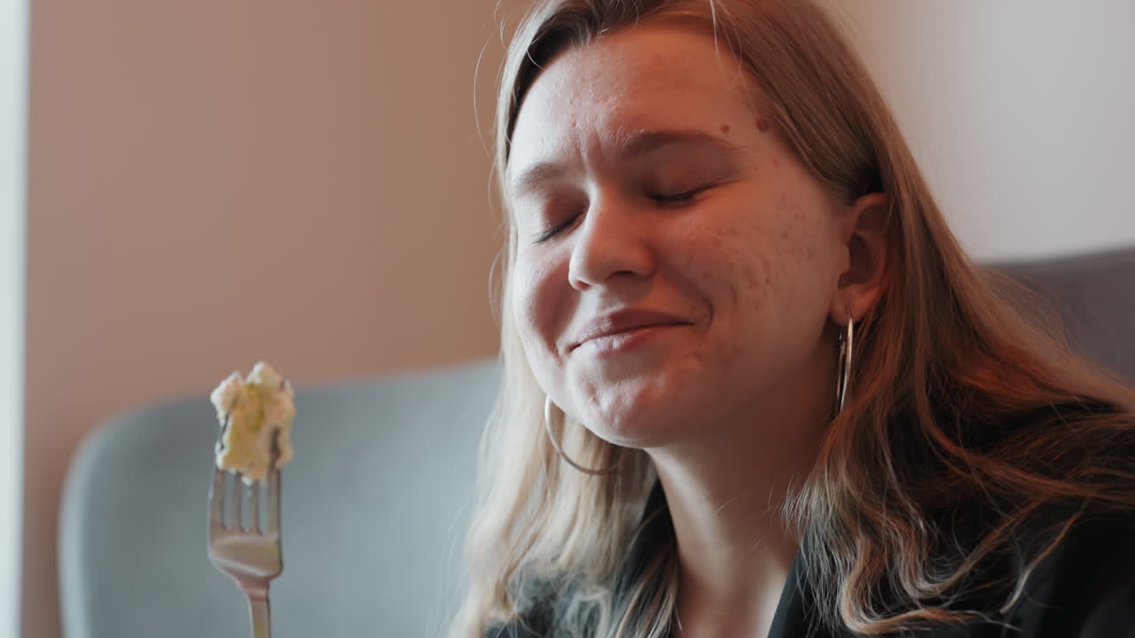 Giovane donna bianca che assapora la pasta sulla forchetta, scena accogliente del soggiorno, sorriso soddisfatto e illuminazione calda e soffusa, pelle lentigginosa e orecchini a cerchio, chef casalinga e food blogger che testa una nuova ricetta con delicatezza