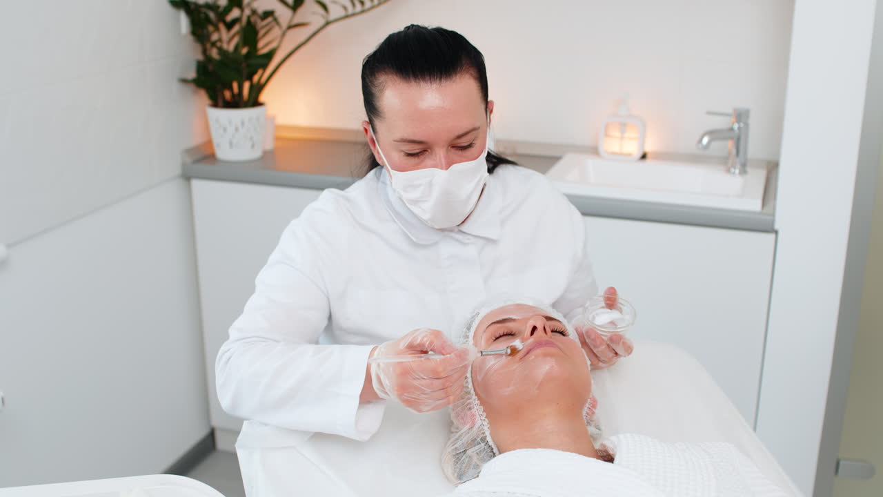 효소 치료 미용사는 청년 성인 여성 얼굴에 브러쉬를 사용하여 보습 의료 크림을 적용합니다.