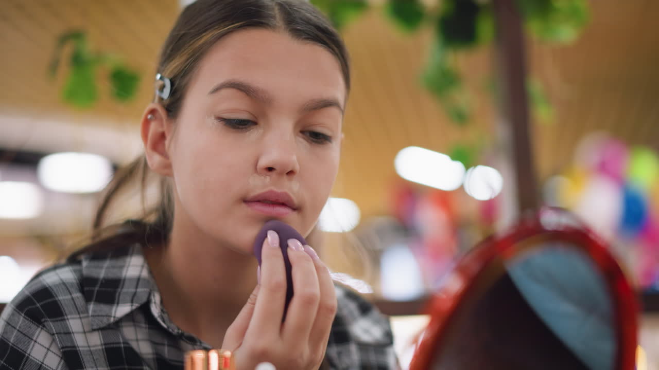젊은 소녀는 부드럽고 자연스러운 마감으로 평평한 피부를 달성하는 데 초점을 맞추고, 따뜻한 조명과 함께 실내 환경에서 화장을 조심스럽게 혼합합니다.