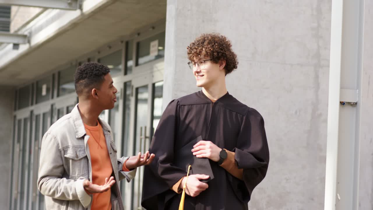 Amigos diversos do sexo masculino caminhando no campus, parabenizando um amigo de beca e capelo que está assentindo.
