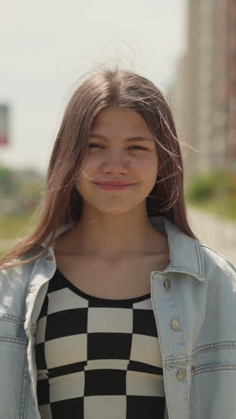 cute girl with long hair smiles looking at camera. teenager wearing denim jacket stands near parked cars on city street on warm day closeup on blurred background