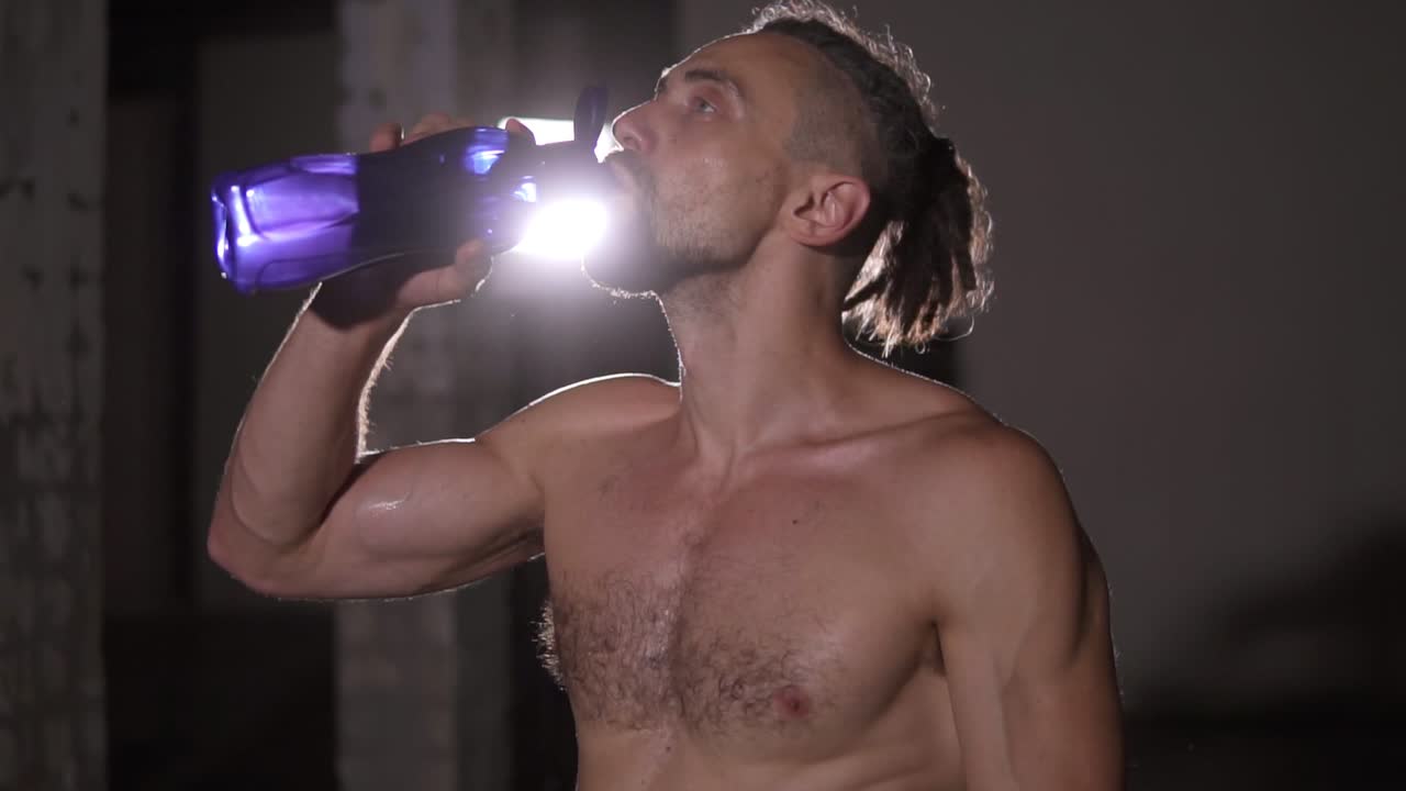 hombre de culturismo con rastas retrato de pie frente a la cámara sin camisa bebiendo sensualmente agua de una botella azul en las luces centro de atención del fondo. cámara lenta