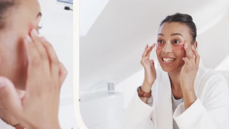 mujer biracial feliz aplicando máscaras debajo de los ojos mirándose en el espejo en un baño soleado, cámara lenta