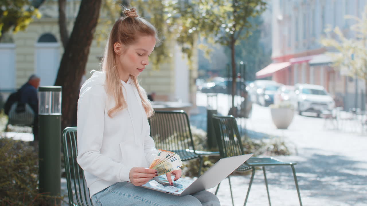 Happy rich woman using laptop winner victory holding cash money in euro banknotes sitting on chair
