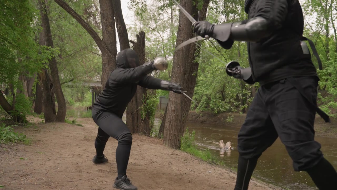 Two men in black fencing gear engage with swords on dirt path surrounded by trees and near river. Their movements are quick demonstrating skill as they parry and thrust. scene is outdoors