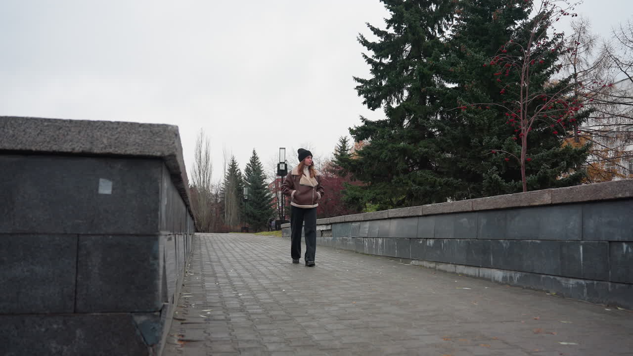 Dreamy girl in black cap brown jacket walking slowly hands in pockets on stone pathway in urban park lined with pine trees and modern buildings under overcast autumn sky