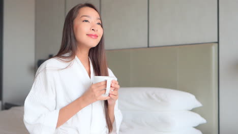 화창한 아침에 침대에서 뜨거운 커피나 차를 들고 목욕 가운을 입은 아름다운 아시아 여성