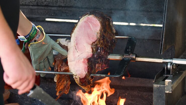 Tangan bersarung tangan mengiris ham babi panggang di atas api terbuka, pencahayaan pasar luar ruangan yang hidup