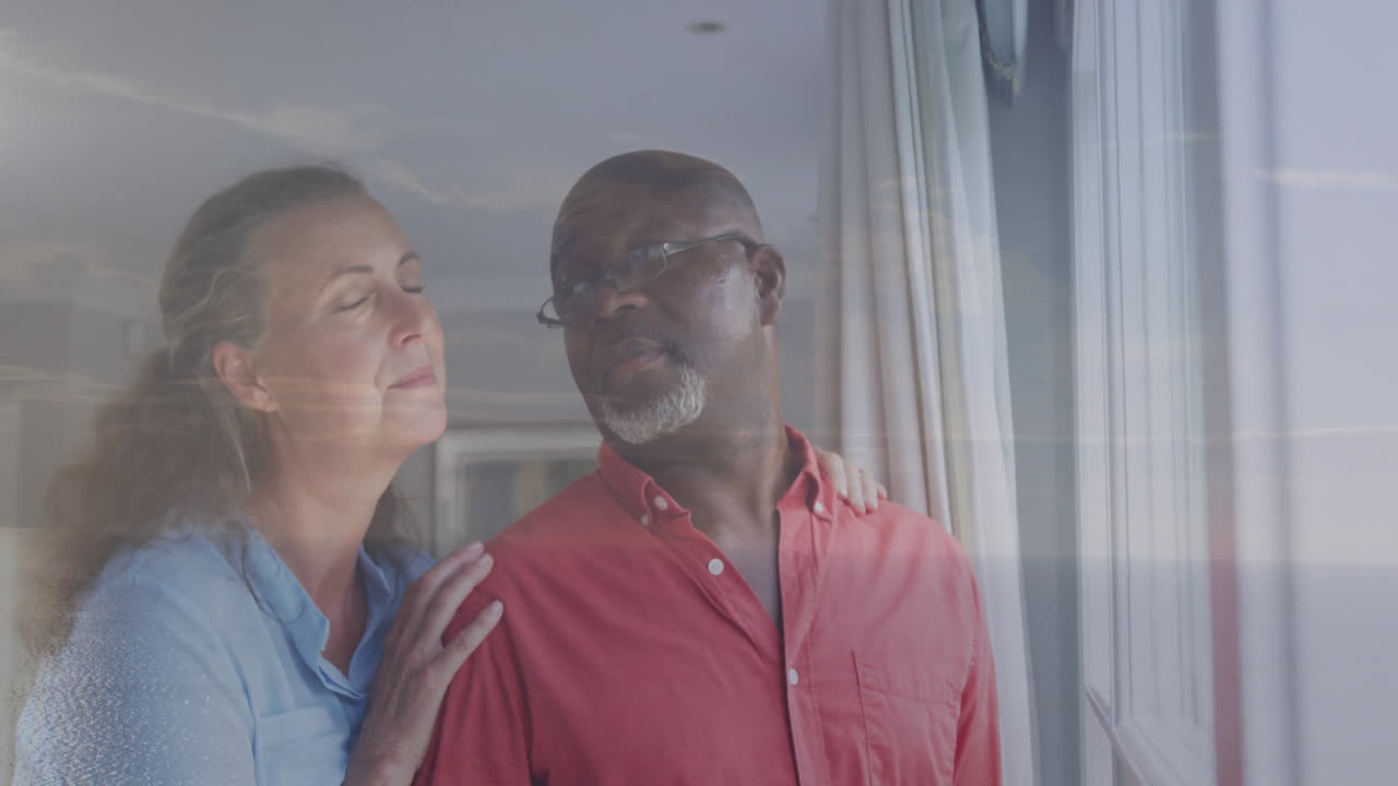 Standing by window, older couple looking outside thoughtfully, sharing quiet moment