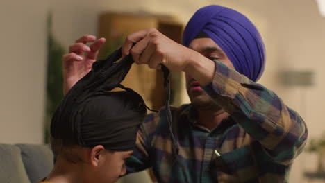 father tying turban onto head of young sikh son with top knot sitting on sofa at home 2