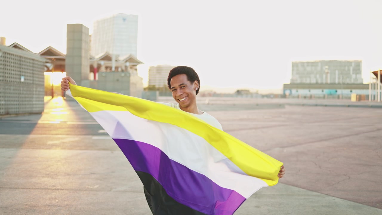 Orang Non-Biner Tersenyum Memegang Bendera Kebanggaan di Luar Ruangan Saat Matahari Terbenam