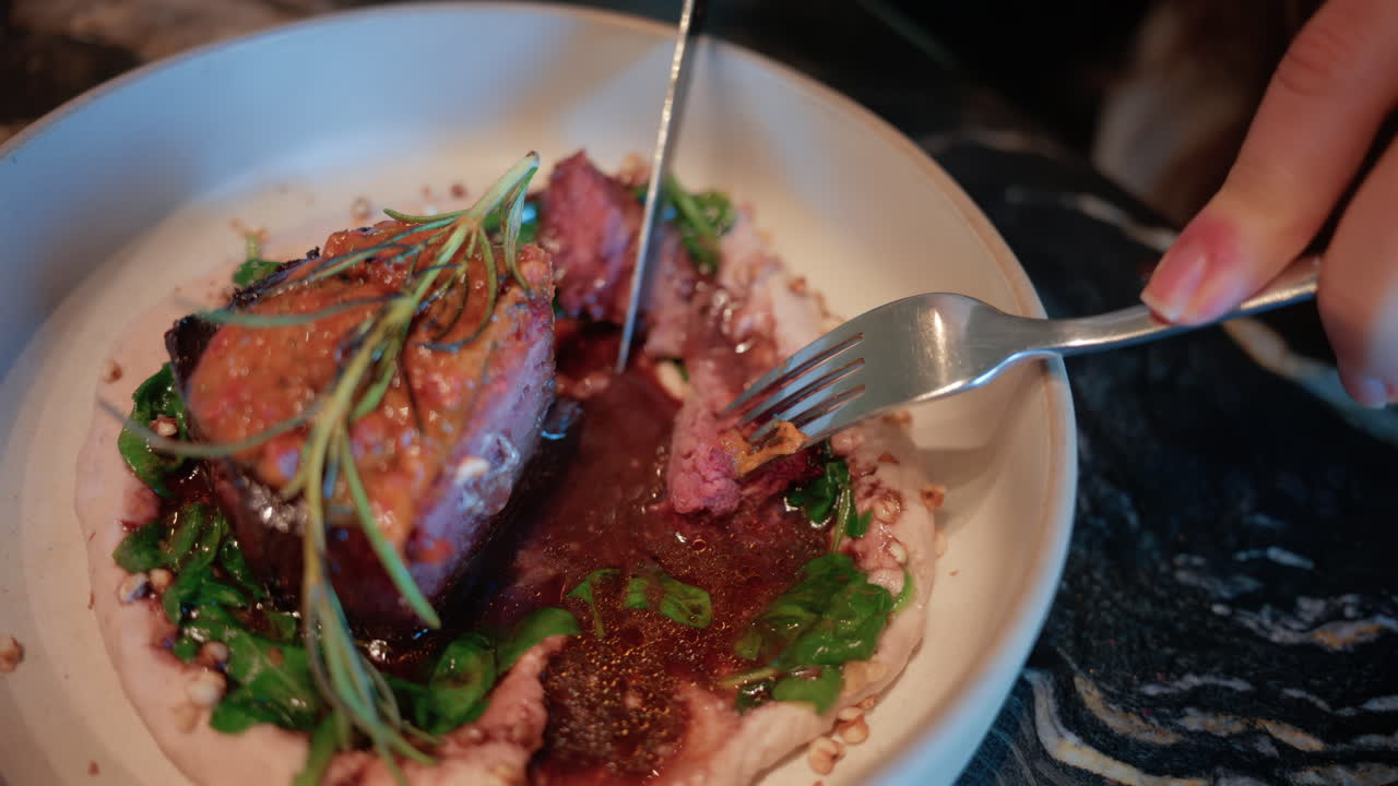 Close up of a woman's hand cutting into a gourmet meat dish served with sauce, greens, and rosemary garnish on a white plate