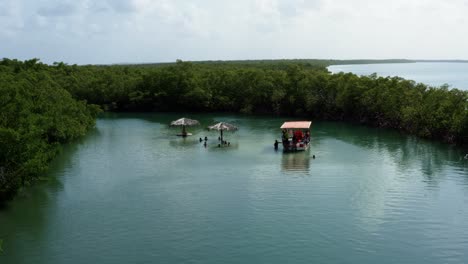 Dolly-in-aerial-drone-shot-of-locals-enjoying-a-small-secluded-natural-turquoise-pool-with-thatch-umbrella's-branching-from-the-Curimataú-river-near-Barra-de-Cunhaú-in-Rio-Grande-do-Norte,-Brazil