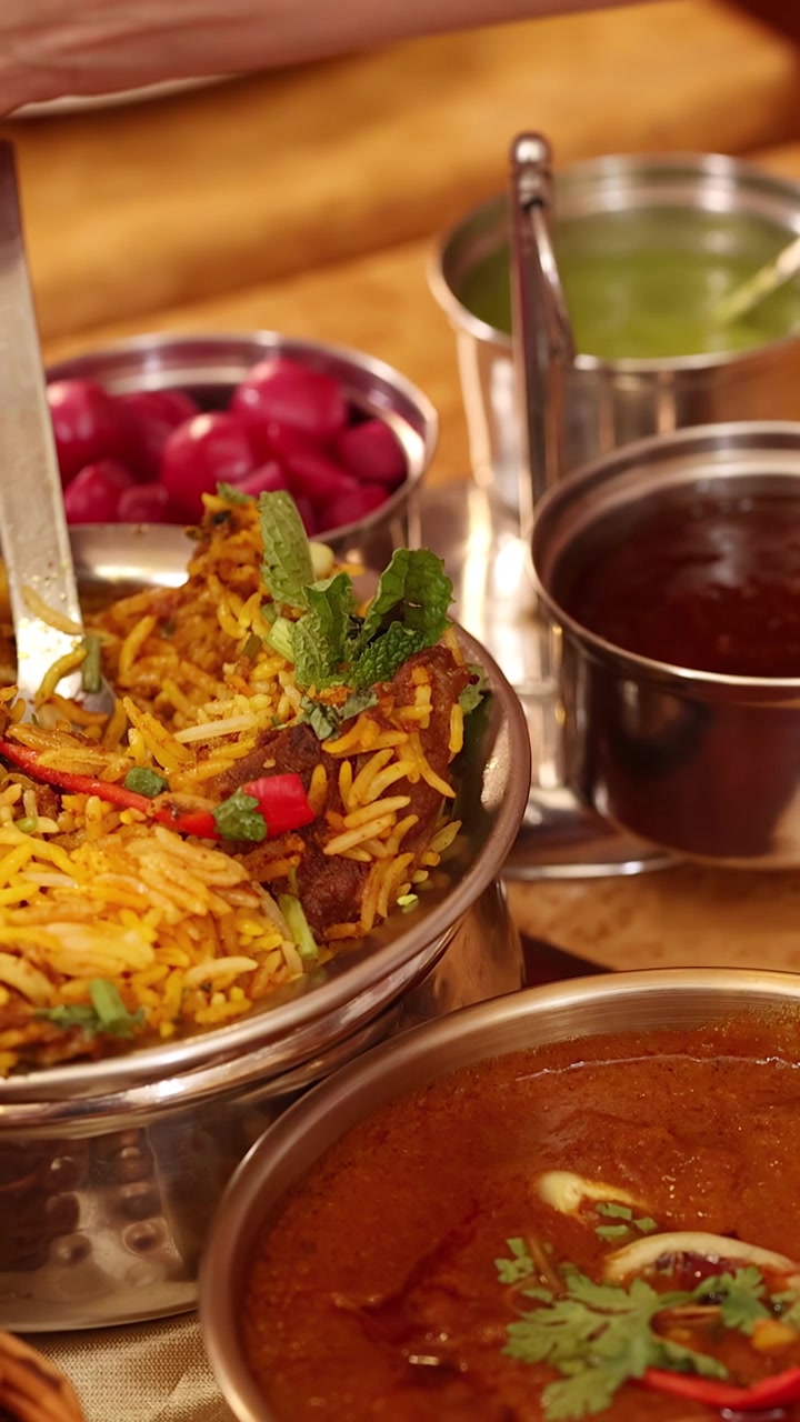Nærbillede af en hånd, der hælder hvid sauce over biryani-ris med forskellige indiske tilbehør.