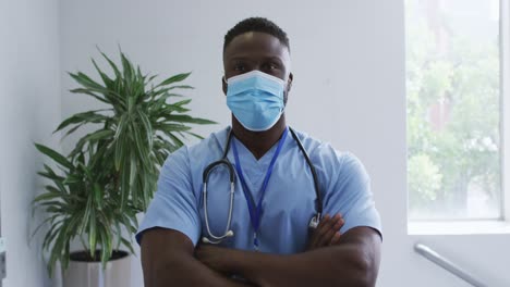 retrato de un médico afroamericano de sexo masculino con bata y máscara facial, con los brazos cruzados en el hospital