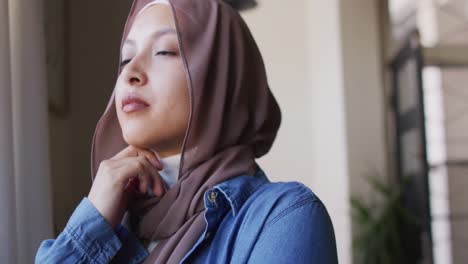 video of thoughtful biracial woman in hijab looking out of window at home