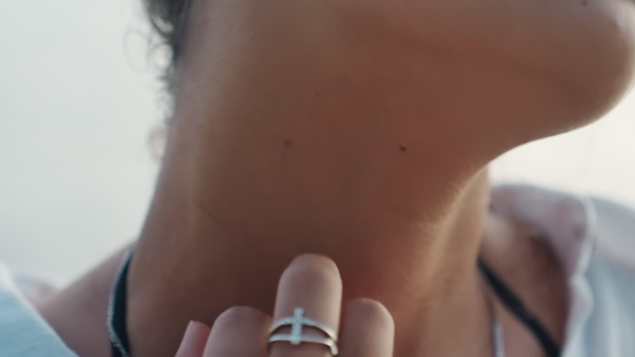 Ritratto ravvicinato del collo con mano che sfiora, anello d'argento al dito, colletto della camicetta e sfondo marino illuminato dal sole, un leggero graffio che suggerisce un'abitudine nervosa, umore pensieroso da viaggiatore, tono cinematografico con messa a fuoco selettiva.