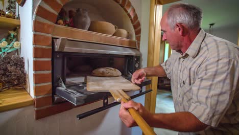 elderly man pulls freshly baked bread out of oven with wooden shovel