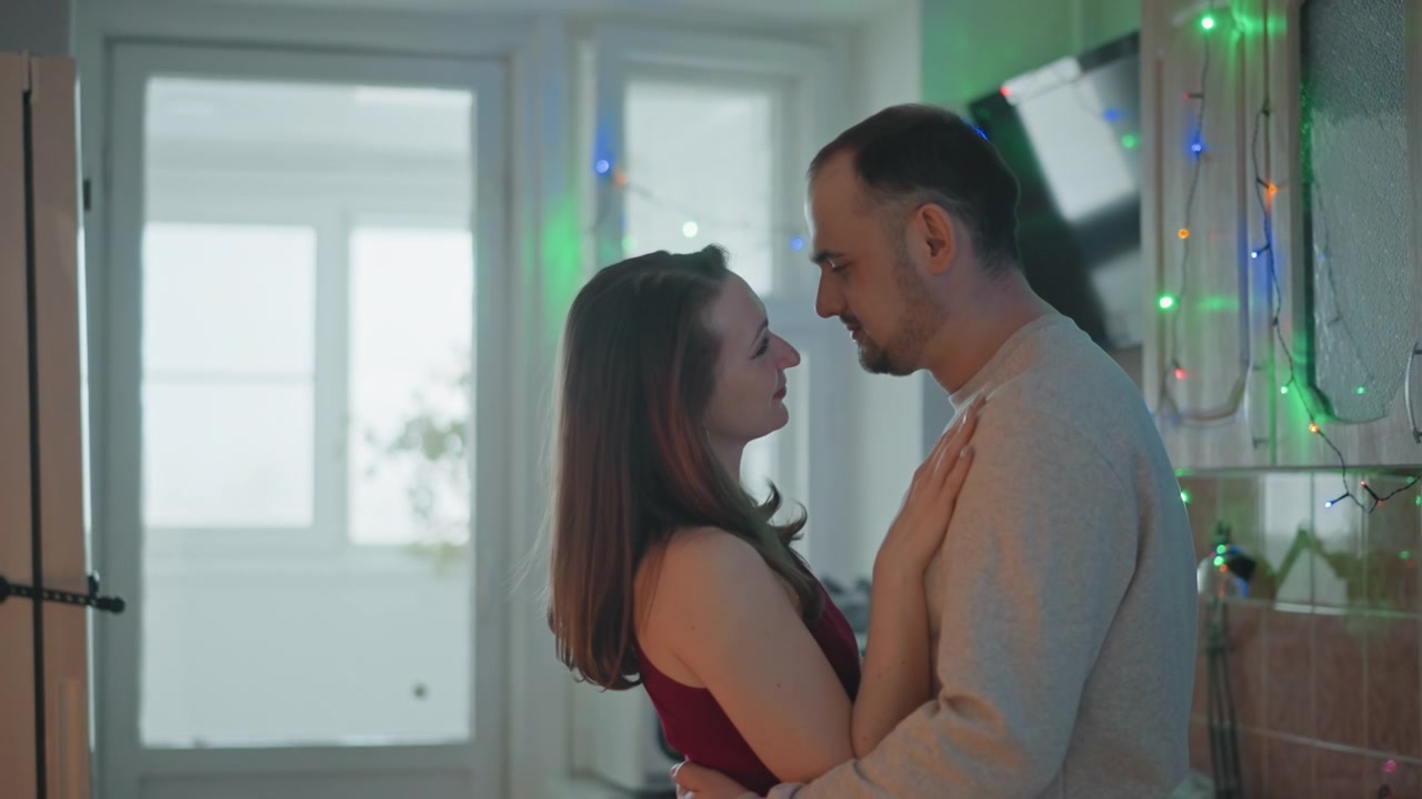 Couple embraces closely inside small kitchen, hands resting at waist and shoulders while leaning together with soft eye contact, slight head tilt, gentle sway, warm window light and cozy domestic mood