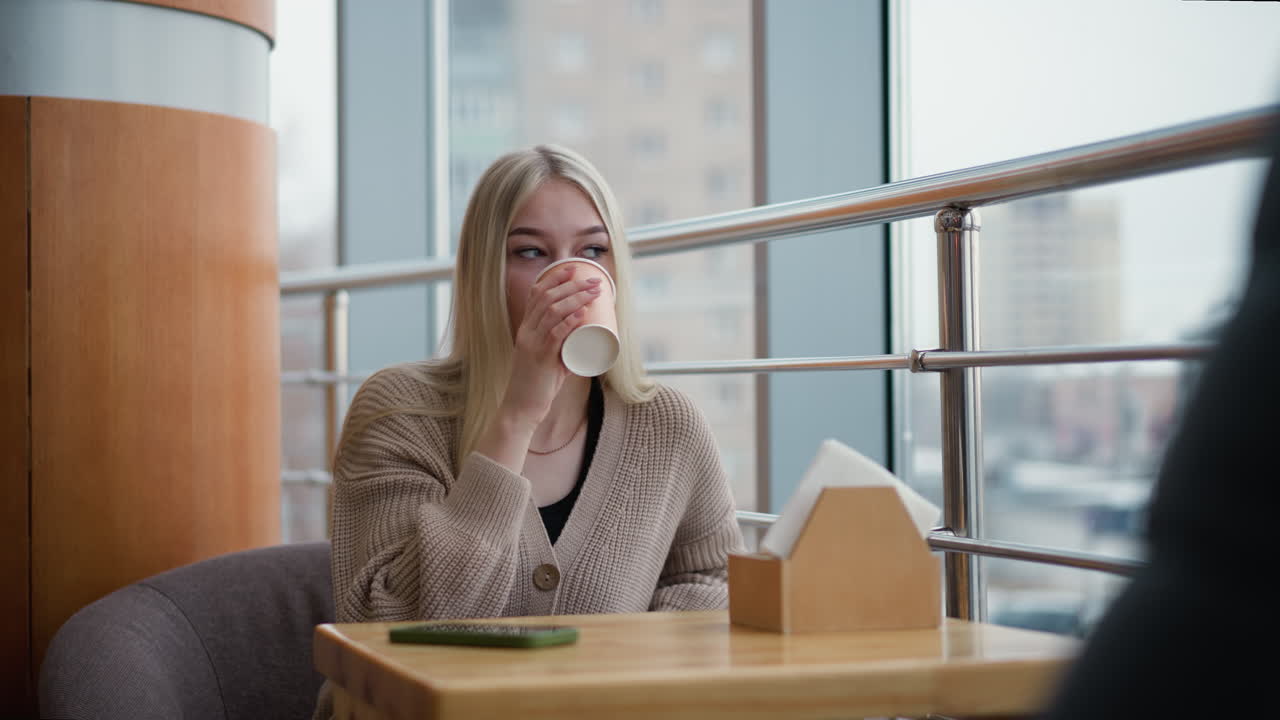 Kvinde sidder og nipper til kaffe i bymiljø med mennesker i bevægelse og biler, der kører forbi i baggrunden, hyggelig indendørsscene med sløret byliv synligt gennem store vinduer.