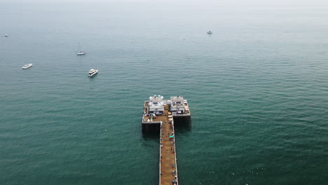 people loafing around on malibu pier with boats littered on the ocean in california usa - forward panning drone shot