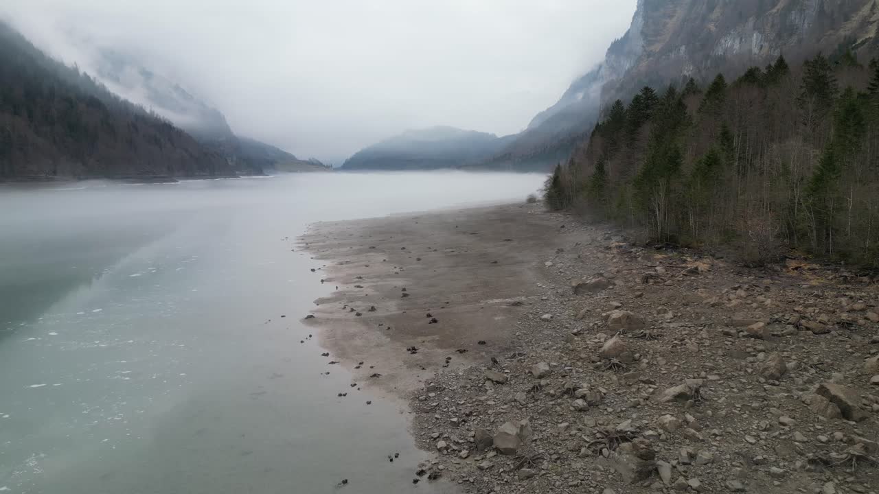 Drone flies along grey frozen shoreline of reservoir lake with misty clouds surrounding mountains in distance