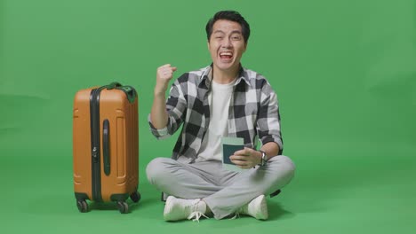 full body of asian male traveler with luggage looking at the passport then screaming goal celebrating while sitting in the green screen background studio