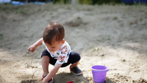 baby boy in light summer clothes takes some sand with his hand. kid stands up, looks at dirty hands and claps them.