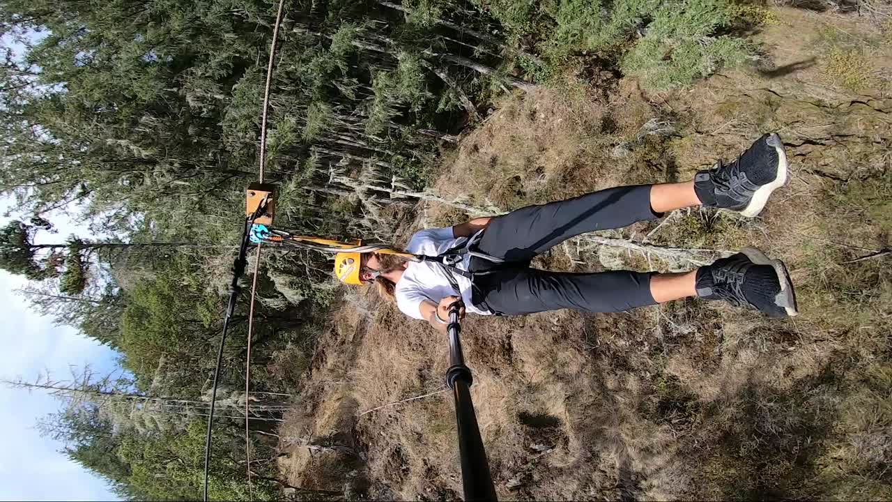 Vertical action camera shot of a young man swinging by very close to trees and bushes in the Canadian forest on the zipline in the hard summer sun on his holiday