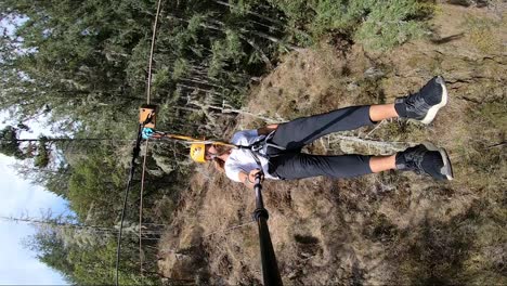 vertical action camera shot of a young man swinging by very close to trees and bushes in the canadian forest on the zipline in the hard summer sun on his holiday