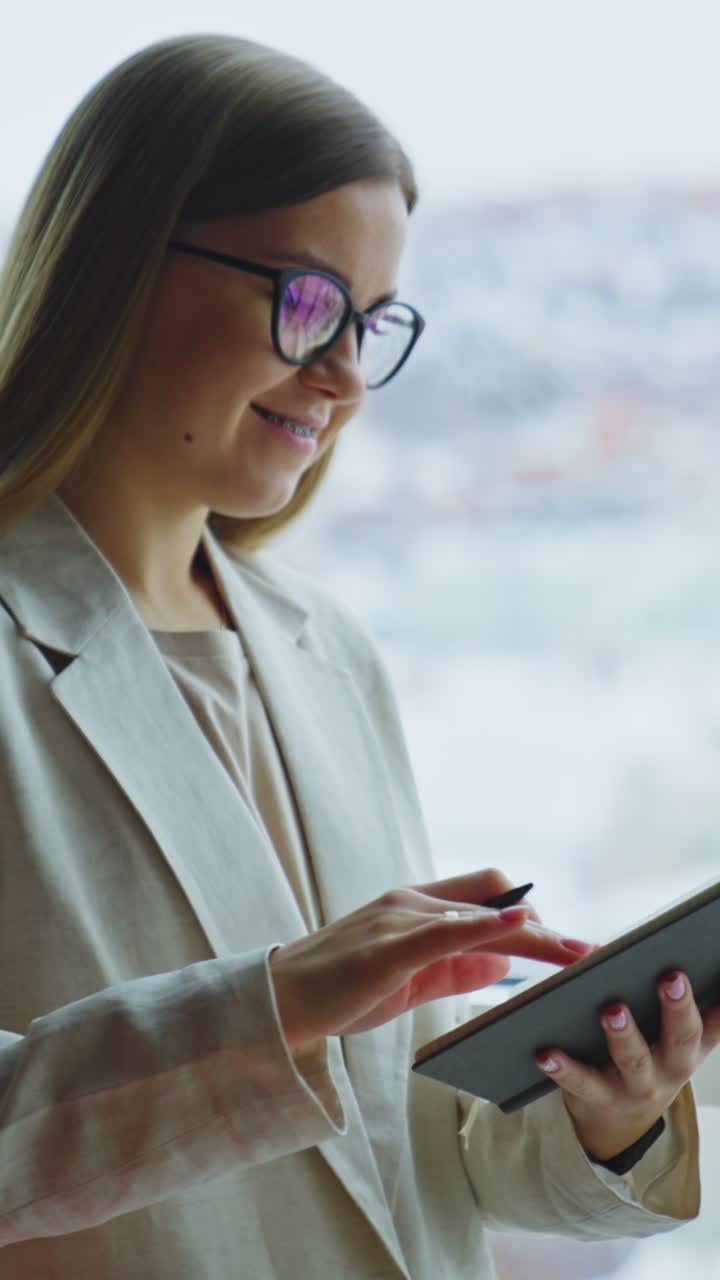 Young woman taking notes in her book standing at the window. Lady in light jacket looks thoughtfully into the blurred window. Vertical video