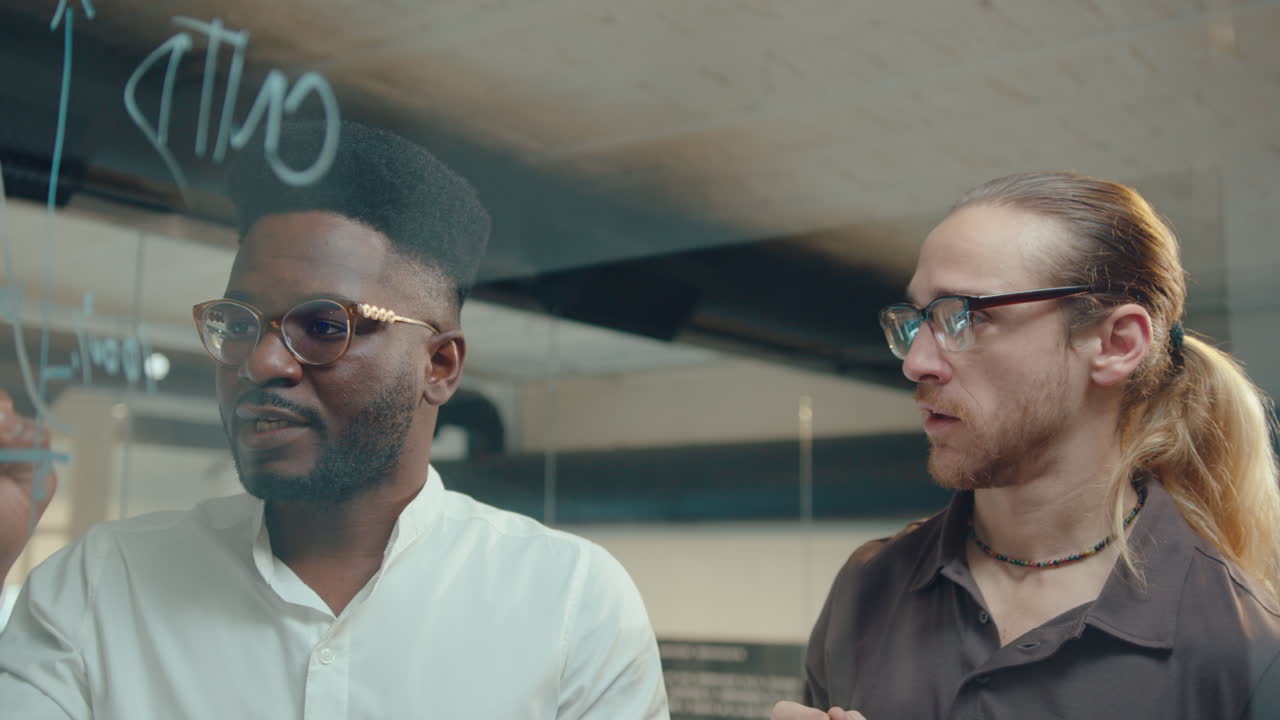 hombres dibujando gráficos financieros en la pared de vidrio y hablando en la oficina