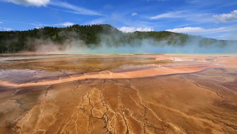 static view of geothermal geyser and hot spring with blue steam cloud rising and mountains in the background at yellowstone national park, wyoming, usa