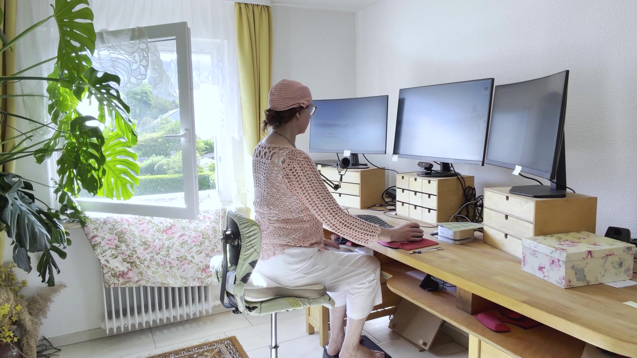 Žena pracuje v ergonomické domácí kanceláři se třemi zakřivenými monitory, ergonomickou klávesnicí a myší a kompaktním stolním počítačem. Statický záběr z úrovně očí.