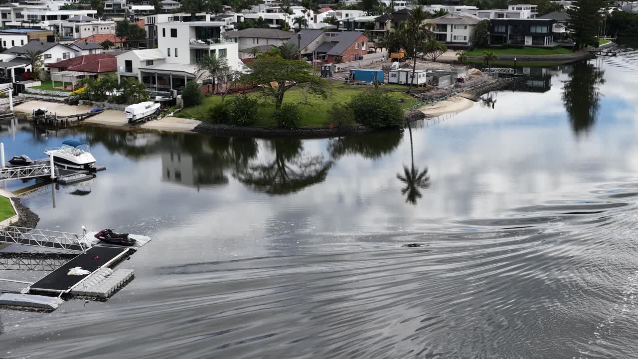 A boat navigates tranquil Gold Coast canals, reflecting luxury homes and palm trees under soft daylight