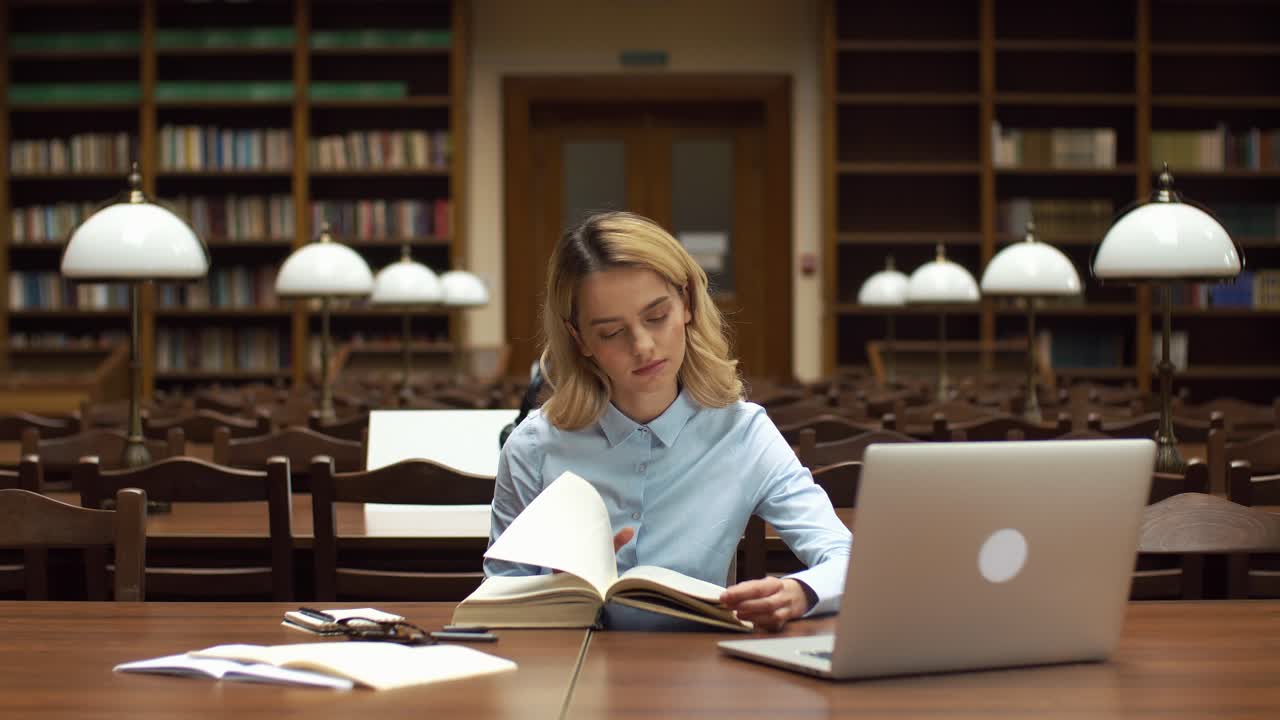 Woman reading a book in a library with a laptop nearby