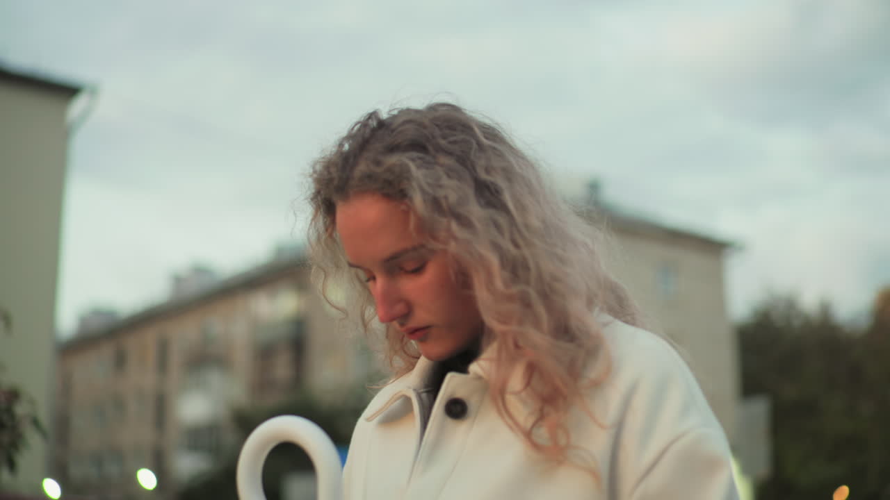 White girl with head bowed reflecting thoughtfully outdoors with blurred view of urban buildings in background during evening light surrounded by calm peaceful atmosphere in city environment