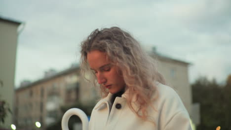 white girl with head bowed reflecting thoughtfully outdoors with blurred view of urban buildings in background during evening light surrounded by calm peaceful atmosphere in city environment