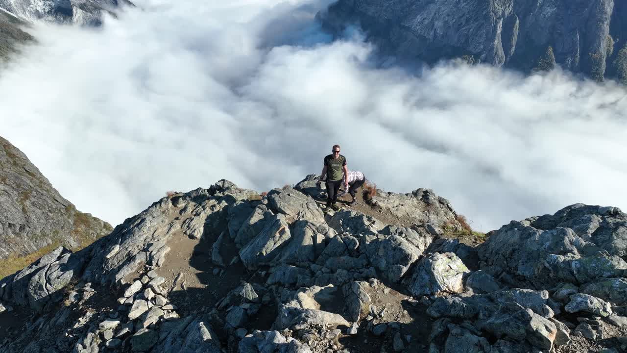 Drone ascends from a couple standing on Bakkanosi cliff, tilting down to reveal the height and clouds covering the fjord far below