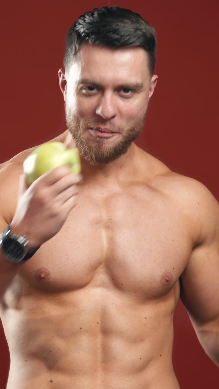 Shirtless bodybuilder eating apple. Portrait of a handsome man with muscular body chooses fresh fruit instead chocolate bar. Smiling strong male eating vitamins in studio. Vertical video