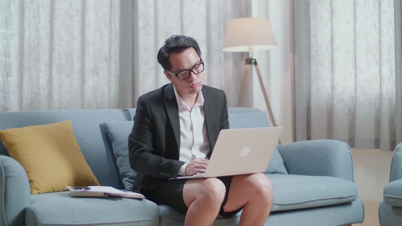 hombre de negocios asiático en chaqueta y pantalones cortos sosteniendo su mejilla y durmiendo mientras trabaja con una computadora portátil en casa
