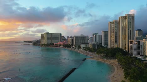 aerial drone view of beachfront resort hotel buildings and people swimming on kuhio beach in waikiki, hawaii at sunset with breakwall