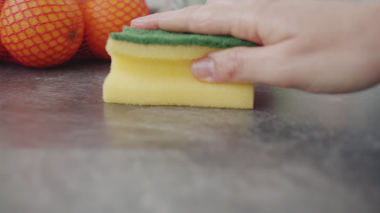 Close up shot of cleaning the kitchen counter with a yellow sponge. Camera dollies back. Slow motion shot with shallow depth of field. Keep the kitchen counter clean