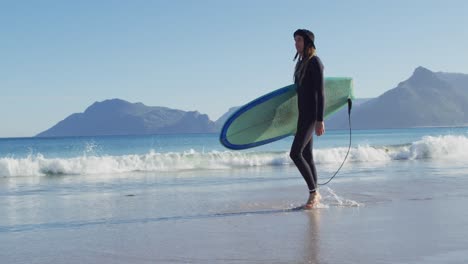 video eines kaukasischen mannes mit dreadlocks in wetsuit, der ein surfbrett trägt und am sonnigen strand spazieren geht