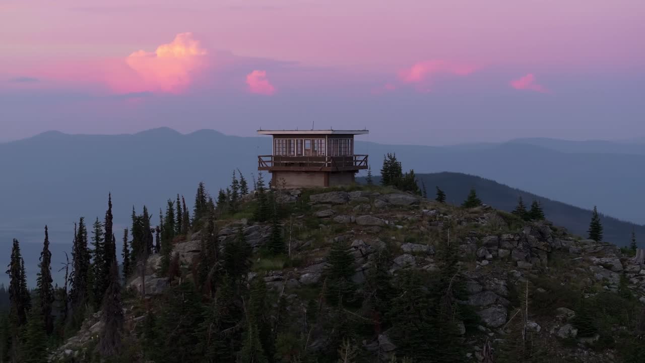 En luftdroneoptagelse, der kredser om et fjerntliggende isoleret brandtårn på en bjergtop i det nordlige Idaho under en smuk farverig solnedgang med en storslået udsigt over bjerge uden mennesker i nærheden.