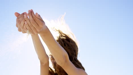 Close-up-of-woman-pouring-sand-running-through-fingers-slow-motion-at-the-beach-with-sun-flare-and-blue-skye