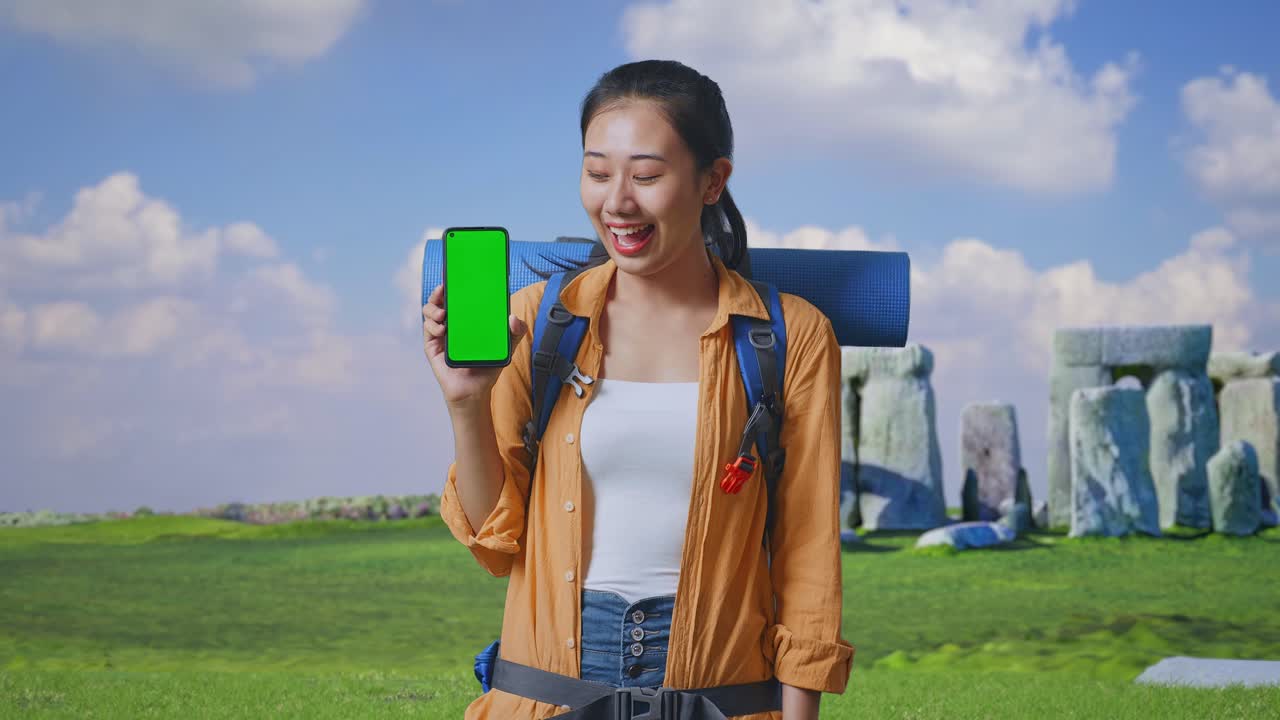 Asian Female Hiker With Mountaineering Backpack Smiling And Showing Mock Up Green Screen Smartphone While Traveling In Stonehenge